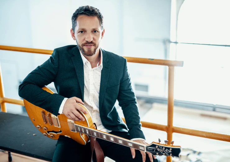 Na barevné fotce je Libor Šmoldas z jazzové kapely Libor Šmoldas Organ Trio, který sedí na lavičce a na klíně má elektronickou hnědou kytaru.